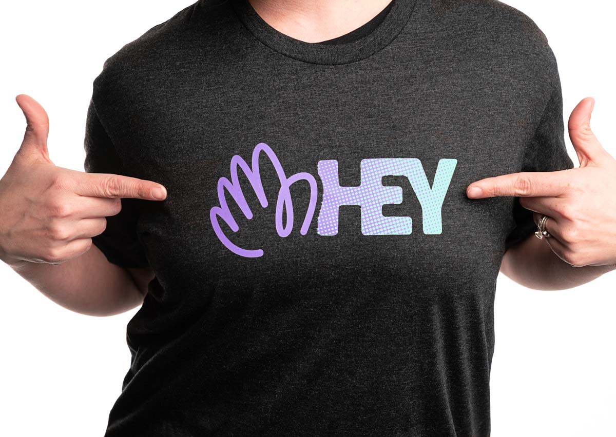 A tightly cropped photo of a person wearing a HEY tee, pointing to the logo on the front of the shirt with both hands